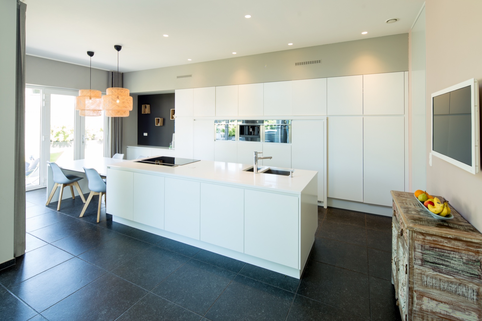 A white custom kitchen with a long island, glowing against a dark tiled floor.